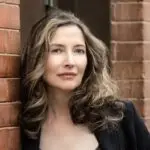 Barbara Nickless, testimonial photo for The Novel View. A woman stands against a rustic brick wall, showcasing a laid-back and confident posture.
